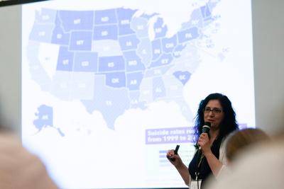 Pictured: A woman with microphone presenting in front of a US map displaying suicide statistics by state