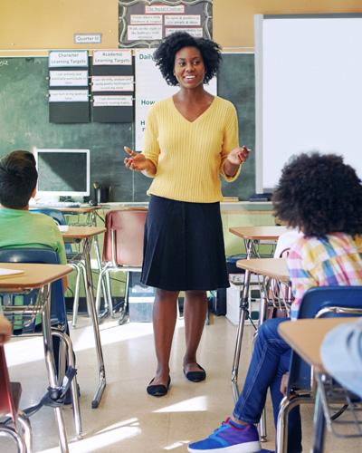 A teacher addresses a classroom of students. Her forearms are extended with palms facing up in a friendly, approchable gesture.